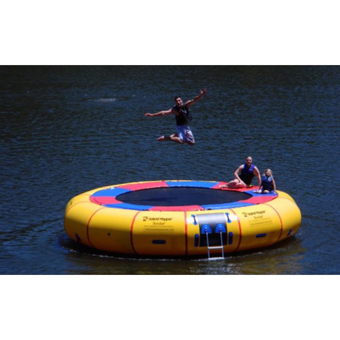 Island Hopper 20ft Acrobat Water Trampoline on a white background with 1 man standing on the black water trampoline surface and 1 girl sitting on the thin red and blue pad that is connected to the yellow inner tube. 