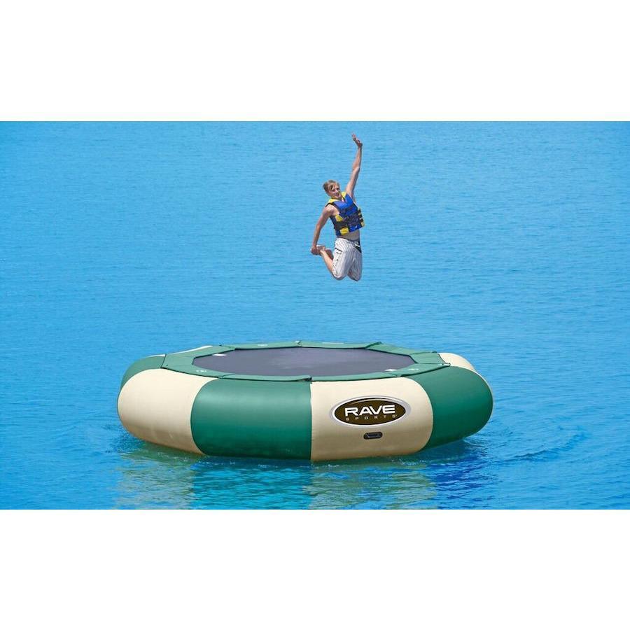 A guy jumping on a green and tan Rave Aqua Jump Eclipse 150 Northwoods Water Trampoline on the lake.