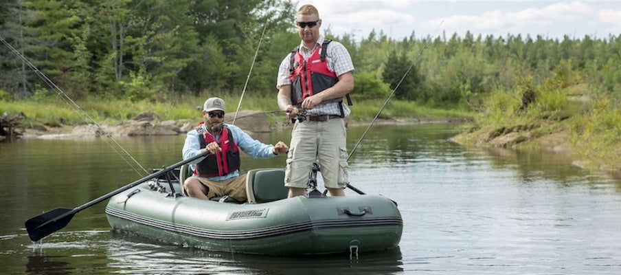 2 men are fishing in a green inflatable boat for fishing. One person is standing up and reeling in a fish. The other is seated and paddling the inflatable fishing boat.