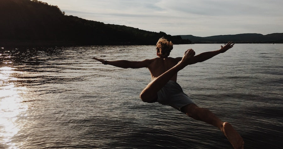 View from behind as a boy jumps into a lake. You can see a big hill filled with trees around the lake.