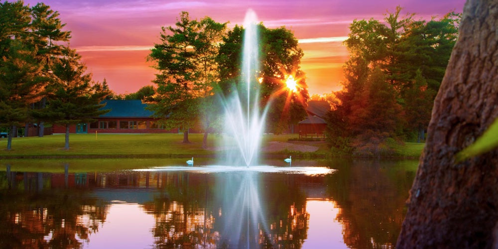 7 Amazing Benefits of a Floating Pond Fountain Blog Photo. A beautiful pond fountain is shown in the middle of the water against a setting sun and pink sky.