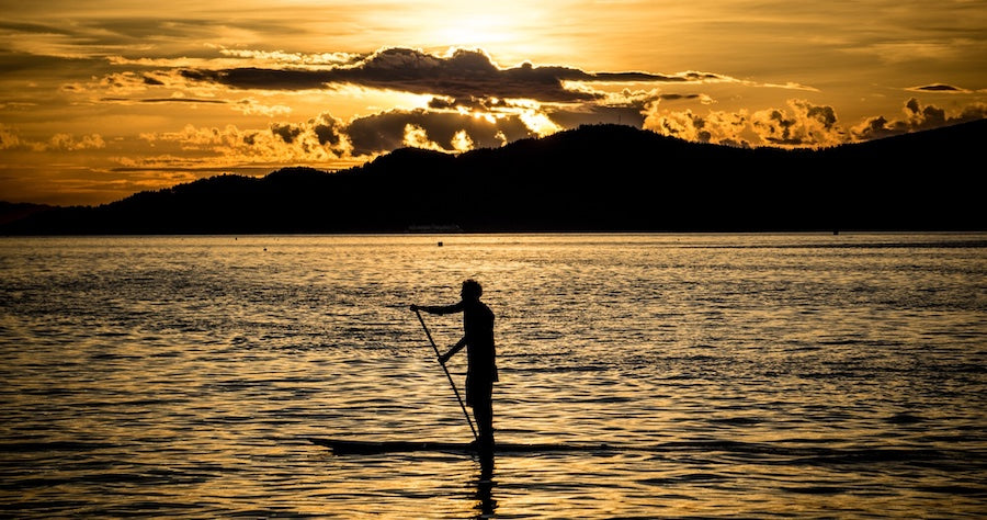 A man paddles on a stand up paddleboard as the sun begins to set behind the clouds over the water. The sky is a blissful mix of orange and blue and is lightly reflected on the water.