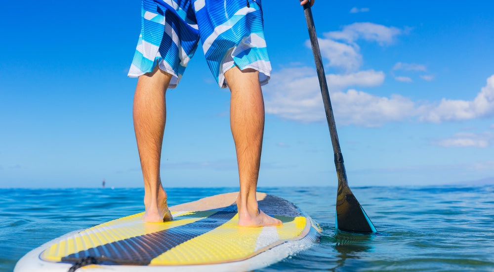 7 Best Watersports for Lake Days. We see a closeup rear view of the legs of a paddle boarder standing on his SUP with a black paddle in hand.
