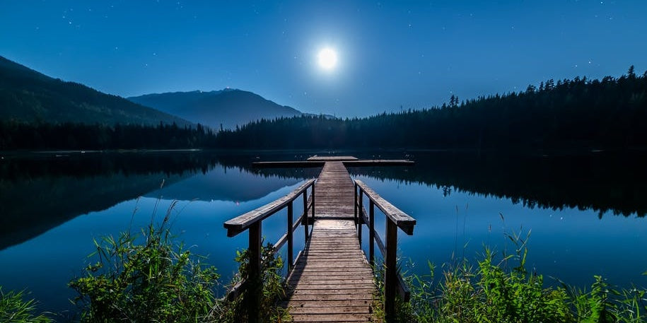 8 Essentials to bring to the lake blog. Shows a dock extending out into a remotely located lake in the mountains. A clear blue sky with bright sun in the middle of the image above the walkway to the dock.