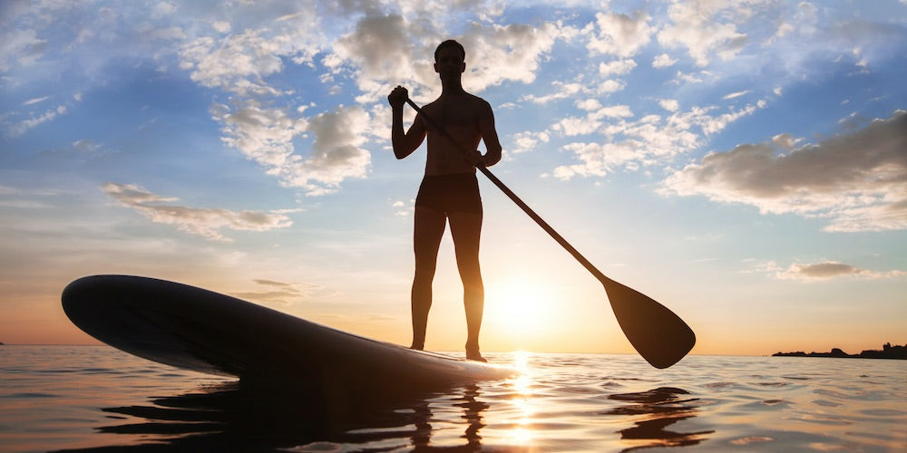 A stand up paddle board is used by a man with the sun behind him.  The sun reflects off the water majestically against the partly cloudy blue sky.