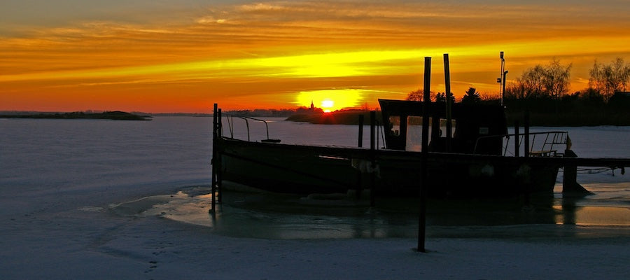 A lake is frozen over, except where an old boat is docked on an old dock. Dock de-icers are keeping the water around the boat from freezing. Beautiful sunset.