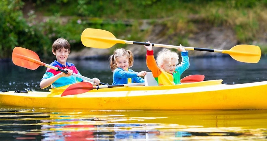 3 kids paddling a yellow kayak. A boy in back has an orange paddle, there is a girl in the middle with no paddle, and a young boy in front has his yellow paddle above his head.