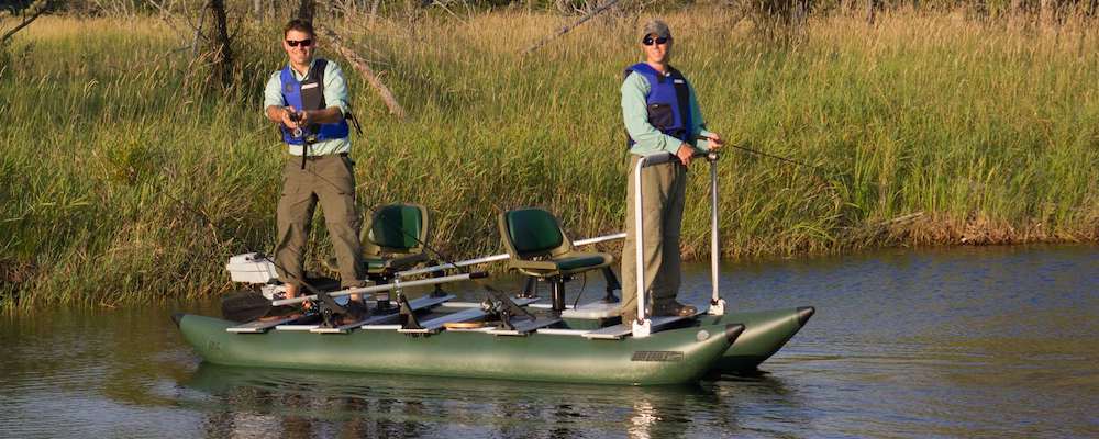 2 men are standing up and fishing on a green inflatable fishing boat. It is a pontoon fishing boat with cross planks and a casting bar. They appear to be near a grassy shore of a smaller lake.