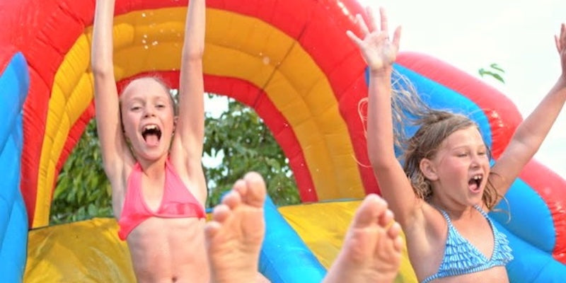 2 young girls sliding down a bounce house with water slide with their hands up. They are having fun!