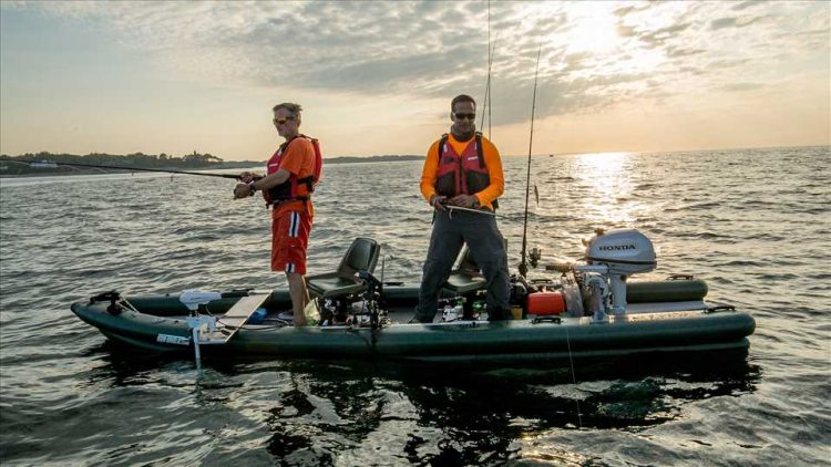 2 men are out on the water fishing in their inflatable fishing boat. They are standing up to fish and everything is calm. They are out on a large lake or ocean with a sunset in the background.