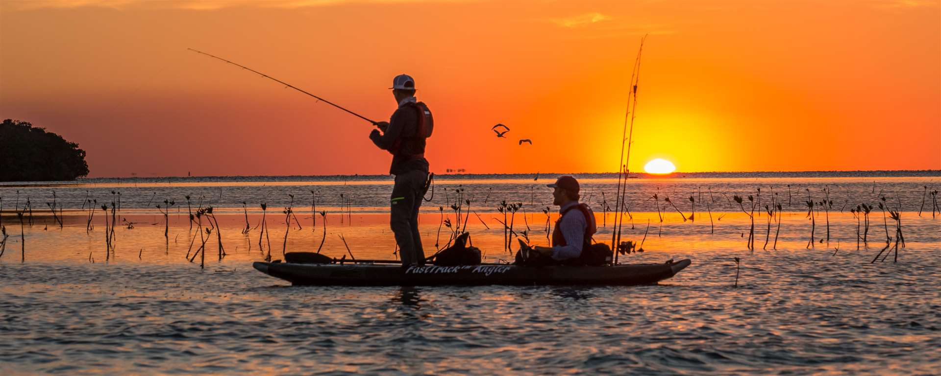 2 men kayak fishing as the sun goes down in an inflatable fishing kayak. The sunsetting on the horizon over the water creates a beautiful orange sky on blue water photo. It is a sit on top fishing kayak, one of the best inflatable fishing kayaks for sale.