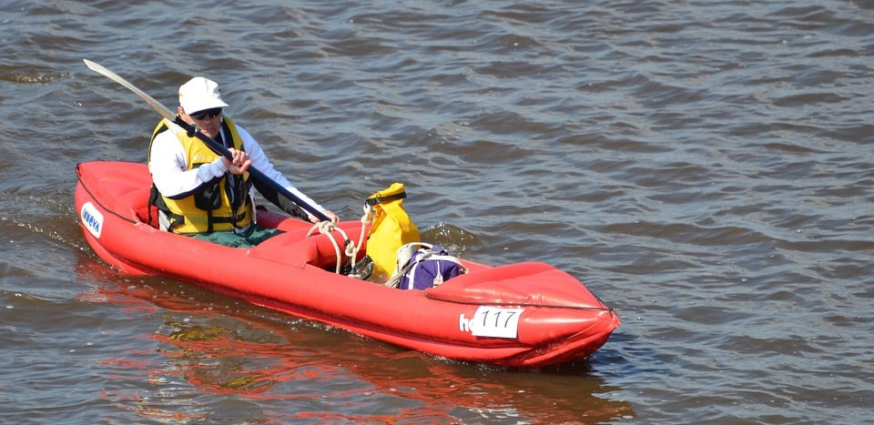 A kayaker paddles his red inflatable kayak on the water. It is a 1 person kayak, or solo kayak, and also could be used as a 2 person inflatable kayak. In this case the front area is being used as storage and has a yellow bag in it.