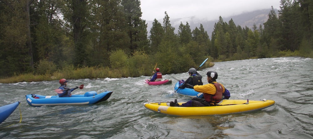 Several people are on a river getting started paddling their inflatable kayak.  There are 4 inflatable kayaks fully pictured, 2 blue, 1 yellow, 1 red.  A fifth inflatable kayak can be seen, but just the front.  It is a very overcast day.