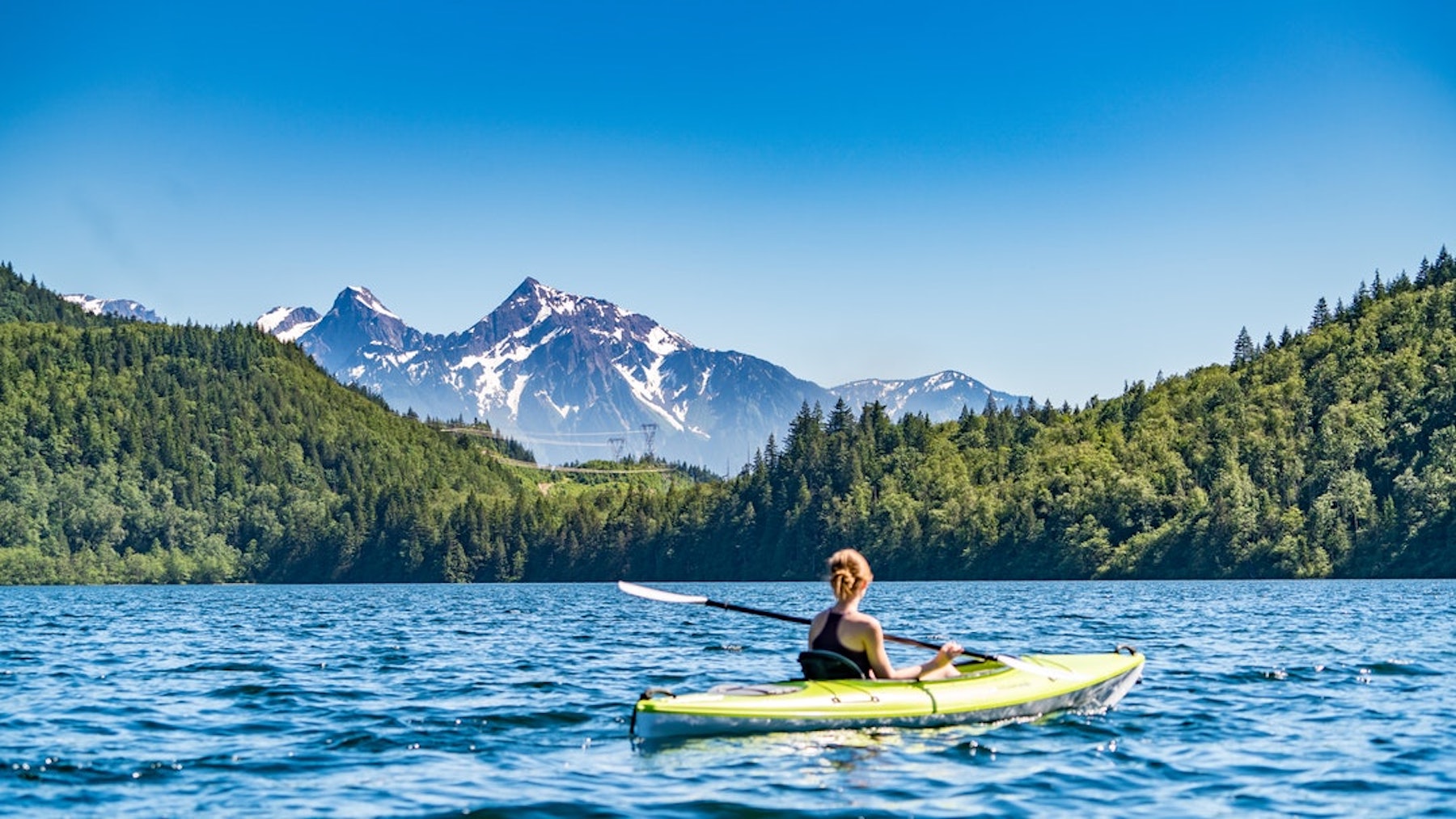 A woman paddles her fishing kayak across the clear blue water. A large mountain is in the background with forestry covering large side hills.