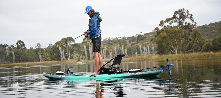 A man stands on a kayak fishing and you can see that he has a kayak motor on the back. He is on a small lake in the fall, as is evident by the trees in the background.