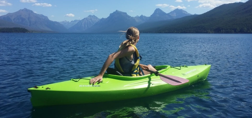 A woman kayaks through Glacier National Park in a green kayak. Mountains in the background. Her kayak is the only watercraft on the water.