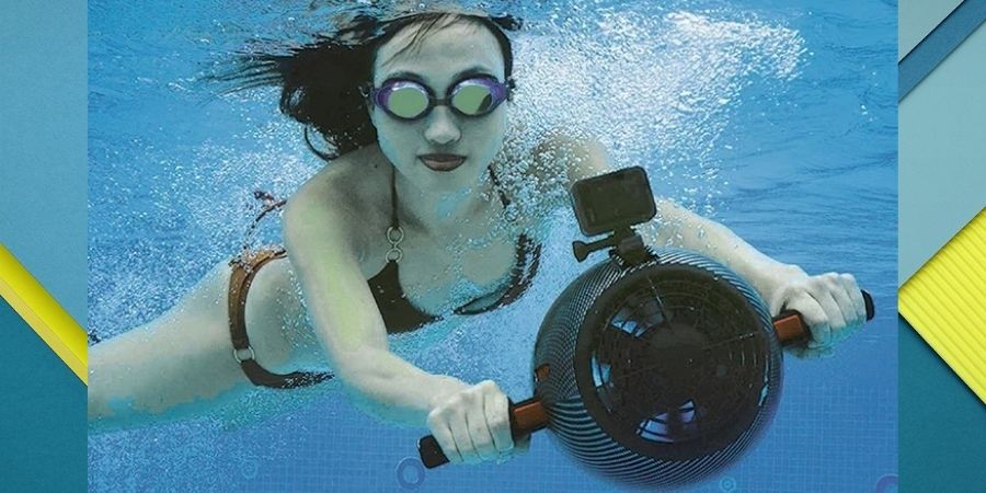 A woman uses a Yamaha Sea Scooter in shallow water. She is in a maroon bikini, the sea scooter is black with a camera on top. She is facing directly at us.