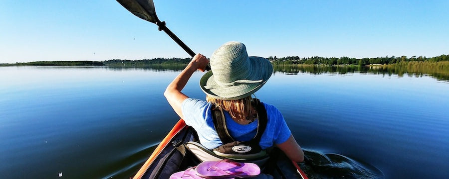 Top Kayaking Tips for Beginners. View from directly behind a kayaker on an open lake. Calm waters, clear skies, and trees wrapped around the lakes edge.