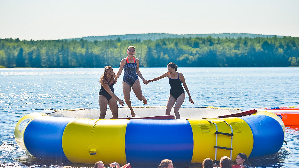 3 young adult ladies jumping on a Rave Water Trampoline on a lake. The yellow and blue alternating panels stick out against the dark blue water and green foliage on a mostly sunny day. 
