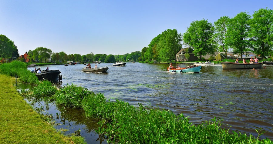 Kayaks, Canoes, and Boat are being paddled down a river. Beautiful green trees on the right and grass on the left of the river.