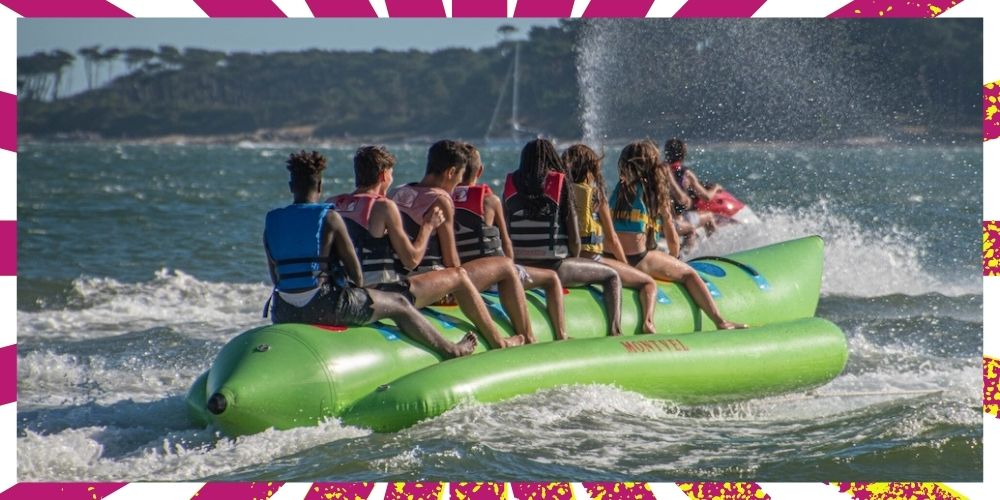 8 kids ride on a green inflatable banana boat tube on a lake. They are pulled by a wave runner.