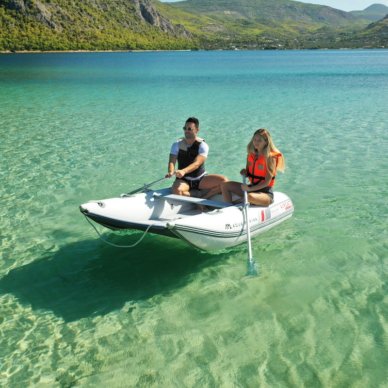 Two people on a small inflatable boat in clear blue water with mountains in the background
