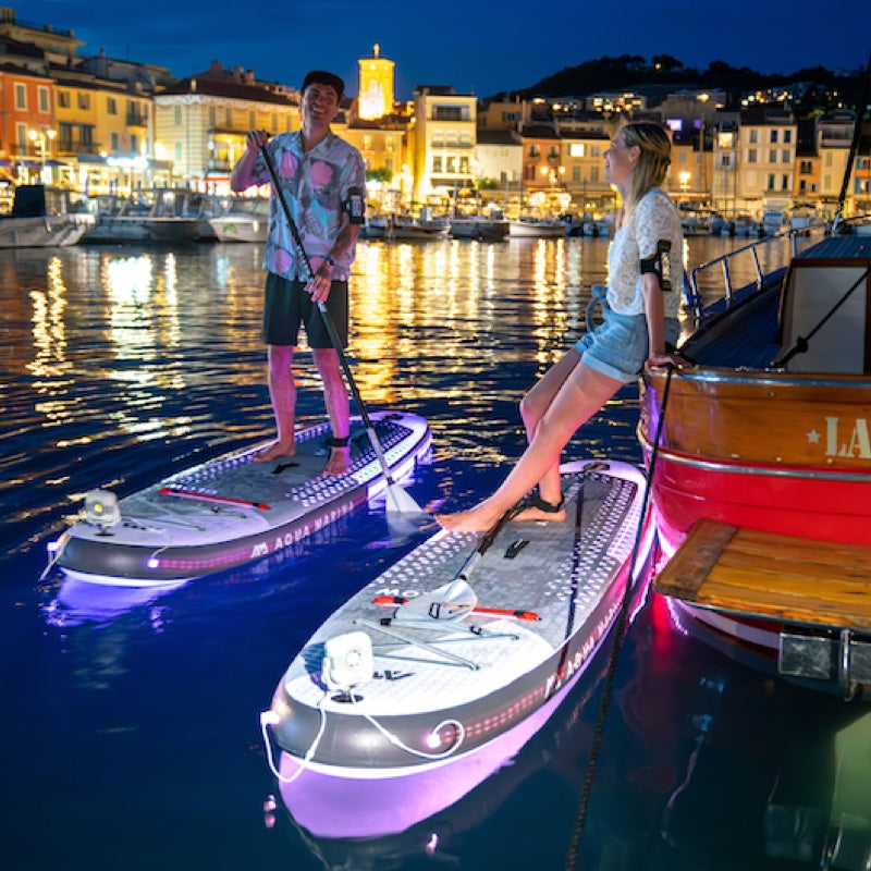 Two people on paddleboards at night in a harbor with illuminated buildings.