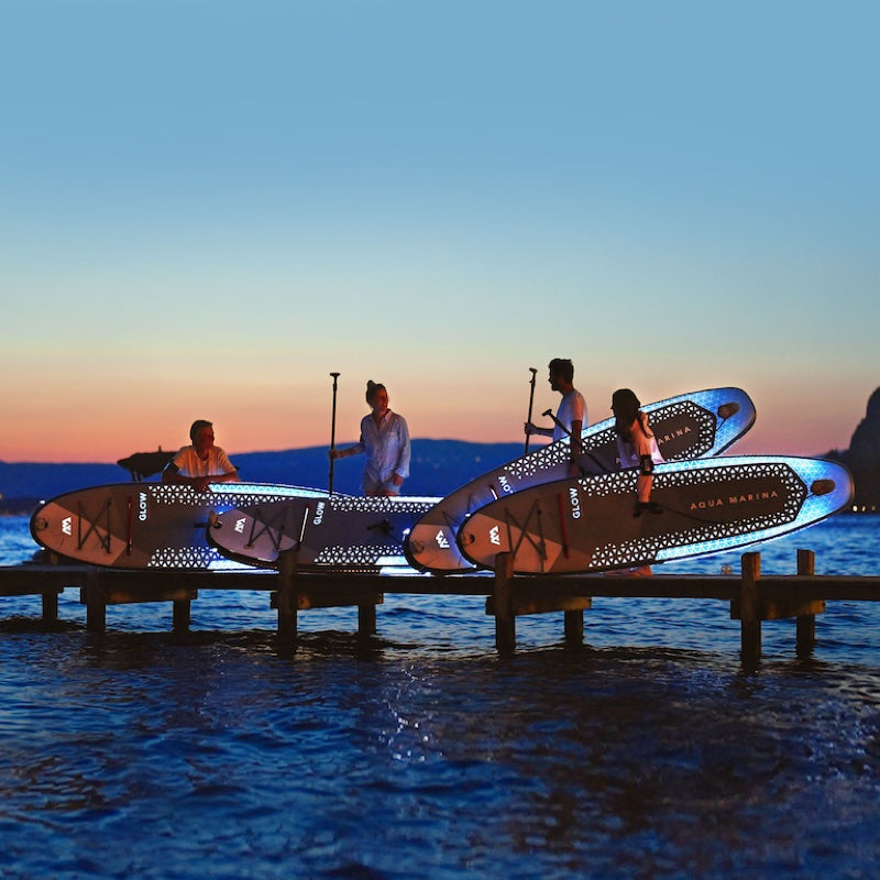 People with illuminated paddleboards on a dock at sunset