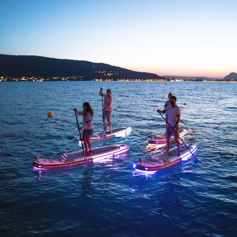 People paddleboarding on illuminated boards in a body of water with a mountainous landscape in the background.