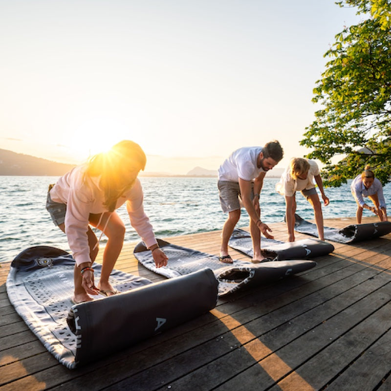 People setting up SUPs on a wooden dock by a lake at sunset.
