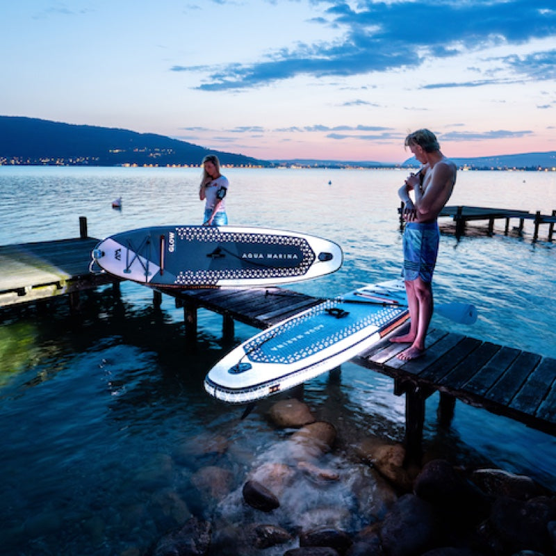 Two people with paddleboards on a dock by a lake at sunset.