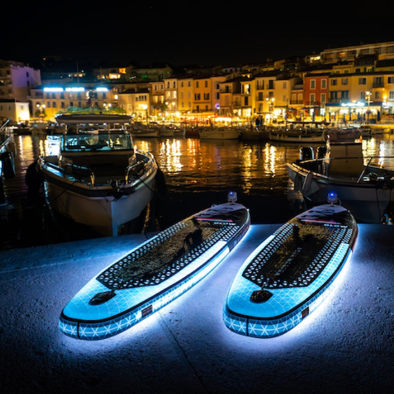 Two illuminated paddle boards on a dock with a cityscape in the background at night.