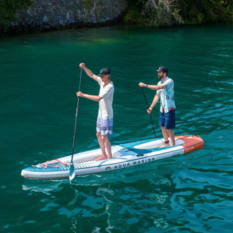 Two people paddleboarding on a clear blue lake with a rocky shore in the background.