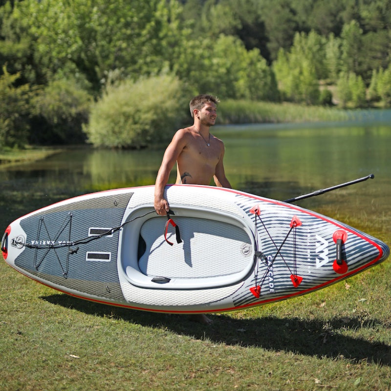 Man carrying an inflatable paddleboard on a grassy area near a body of water with trees in the background.