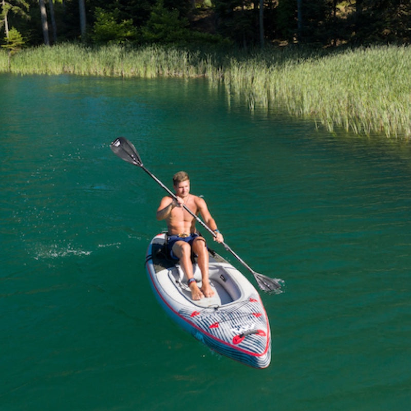 Man paddling a kayak on a clear lake with trees and reeds in the background