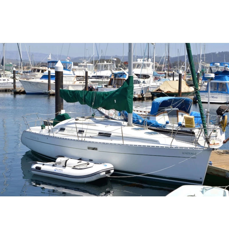 White sailboat with a green sail and white dinghy at a dock with other boats in the background.