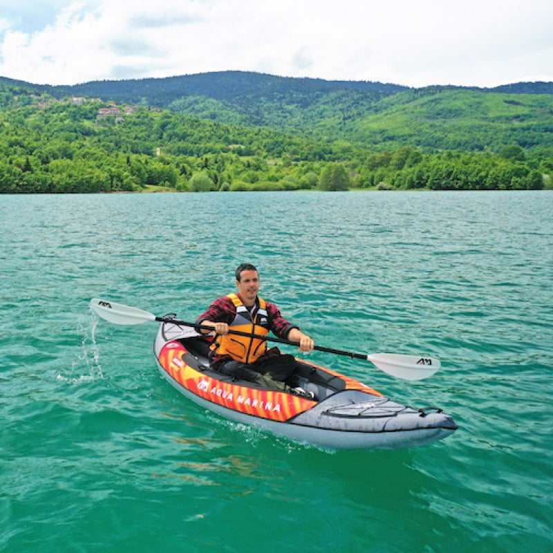 Man paddling an inflatable kayak on a calm lake with green mountains in the background