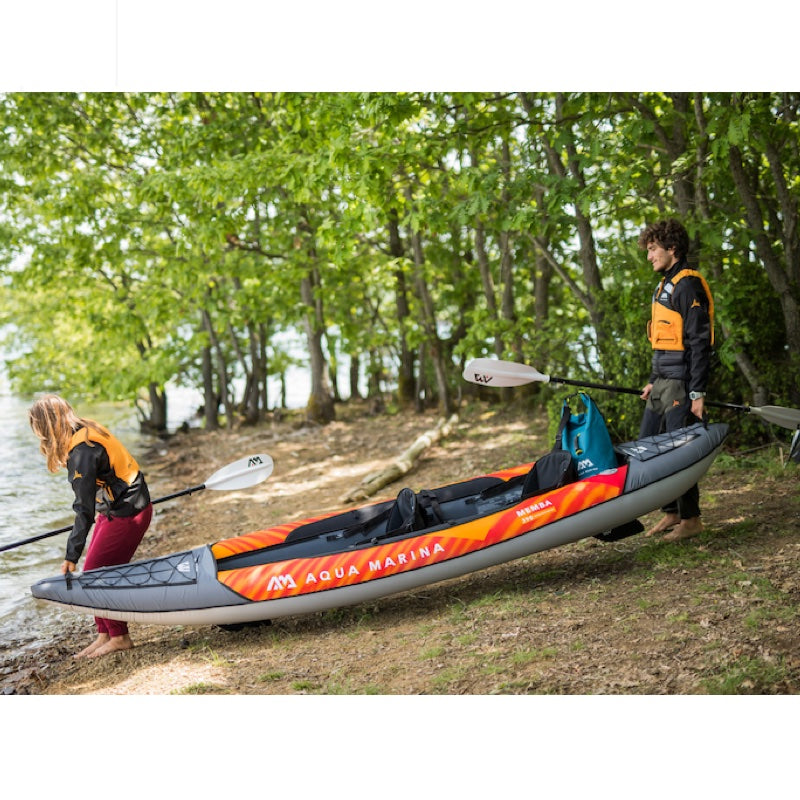 Two people with an Aqua Marina kayak on a lakeside path with trees in the background.