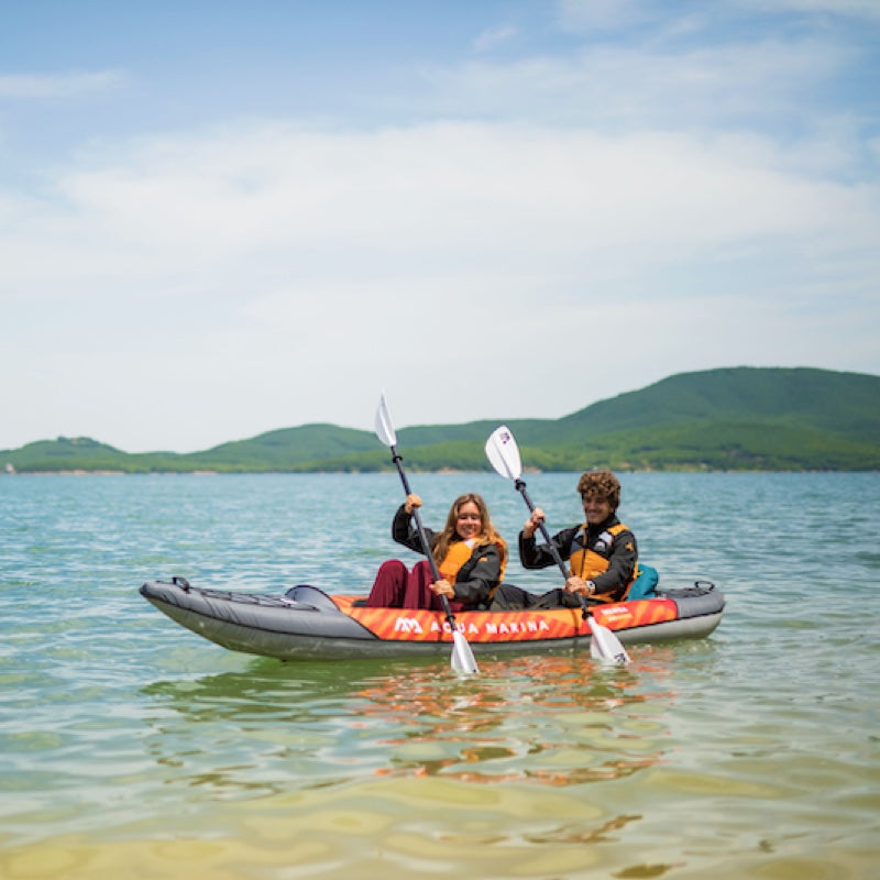 Two people kayaking on a calm lake with mountains in the background