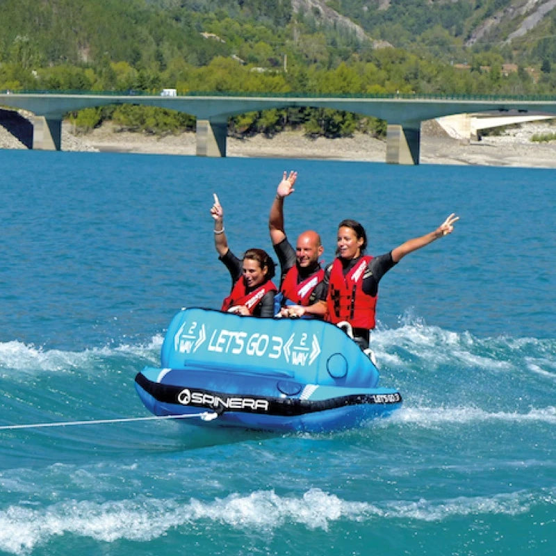 Three people on a blue inflatable tube with 'Spinera' branding in a body of water with a bridge and greenery in the background.