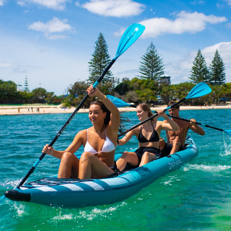 Three people in an aqua kayak paddling on clear blue water with trees and beach in the background.