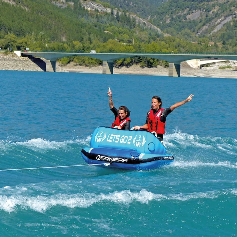 Two people on a blue inflatable tube in a lake with a bridge and mountains in the background.