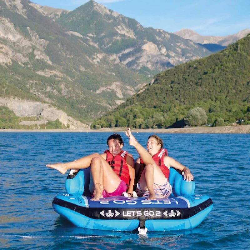 Two people on a blue inflatable tube in a lake with mountains in the background