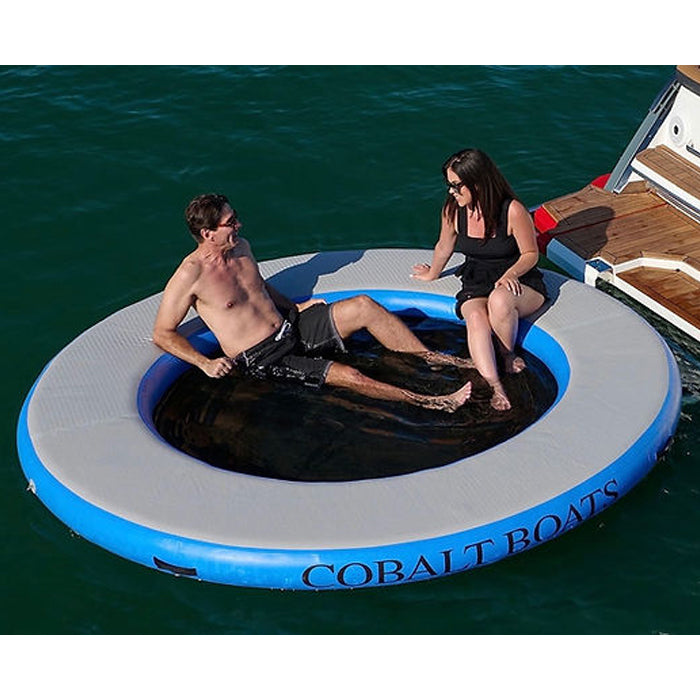 The blue variation of Paradise Pad 8ft Splash Island Water Mat with 2 people hanging out on it by the boat.