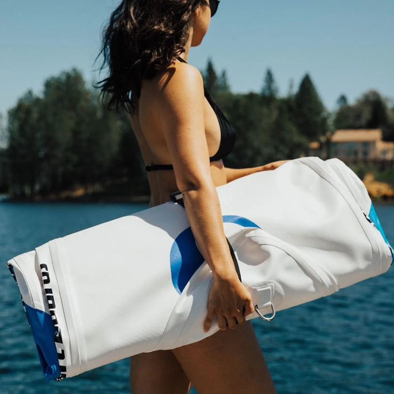 Person holding a white and blue towel by a body of water with trees and a house in the background.