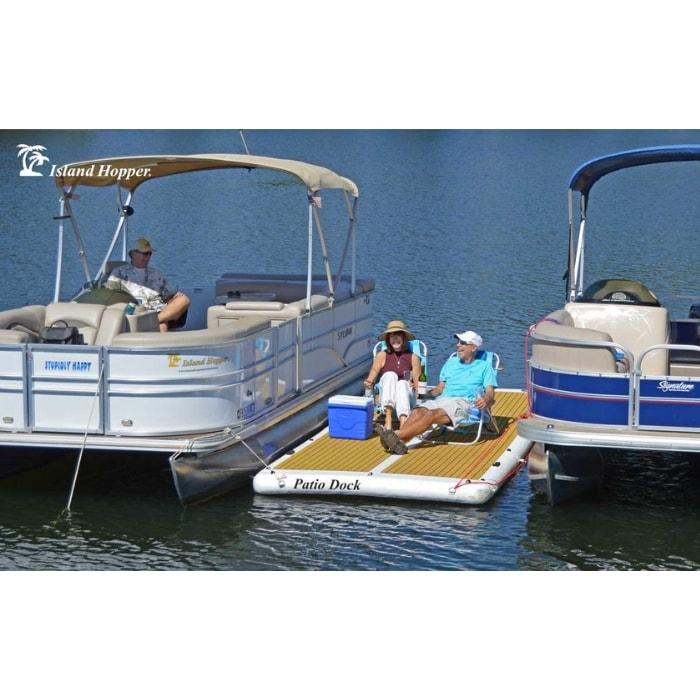 The Island Hopper Patio Dock Floating Swim Platform is shown here attached in between 2 pontoon boats. There are 2 people sitting in chairs on the inflatable floating dock along with a cooler. The water is dark blue and is visible in front and behind the inflatable dock and the boats.