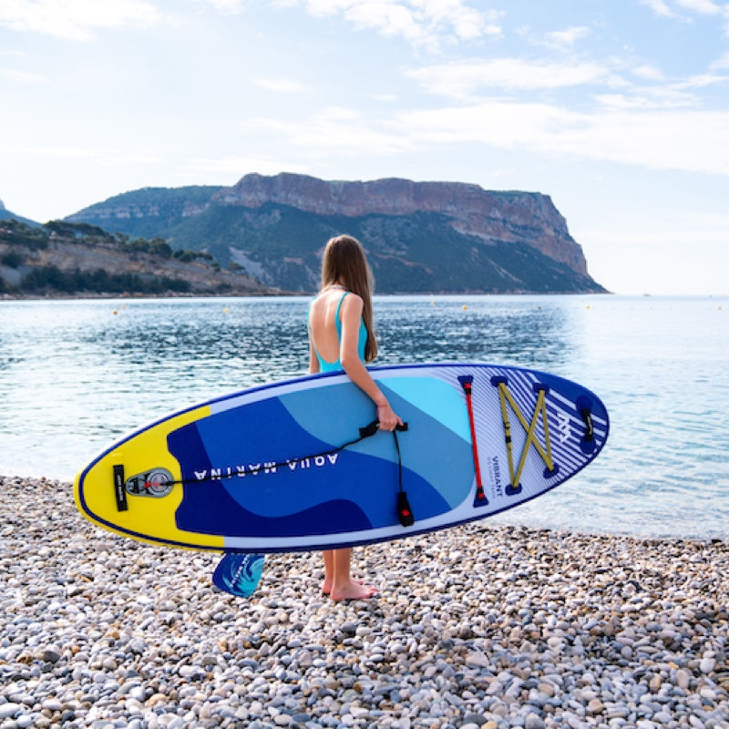 Woman holding a blue and yellow paddleboard on a pebbly beach with mountains in the background