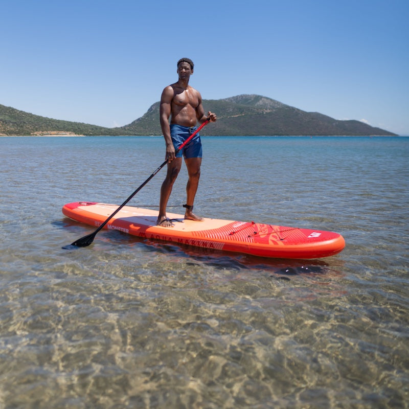 Man standing on a red paddleboard in clear water with mountains in the background