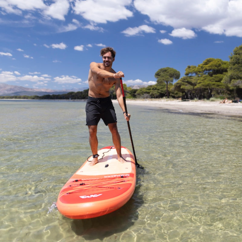 Man paddleboarding on a clear body of water with a beach and trees in the background