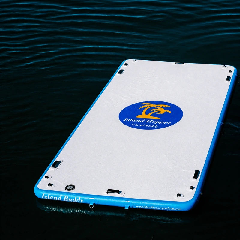Island Hopper 12ft Island Buddy Inflatable Water Mat sitting in the middle of the lake by itself. It is white on top with a royal blue oval. Inside the oval is yellow palm trees and white Island Hopper Island Buddy lettering in the oval and on the sides.